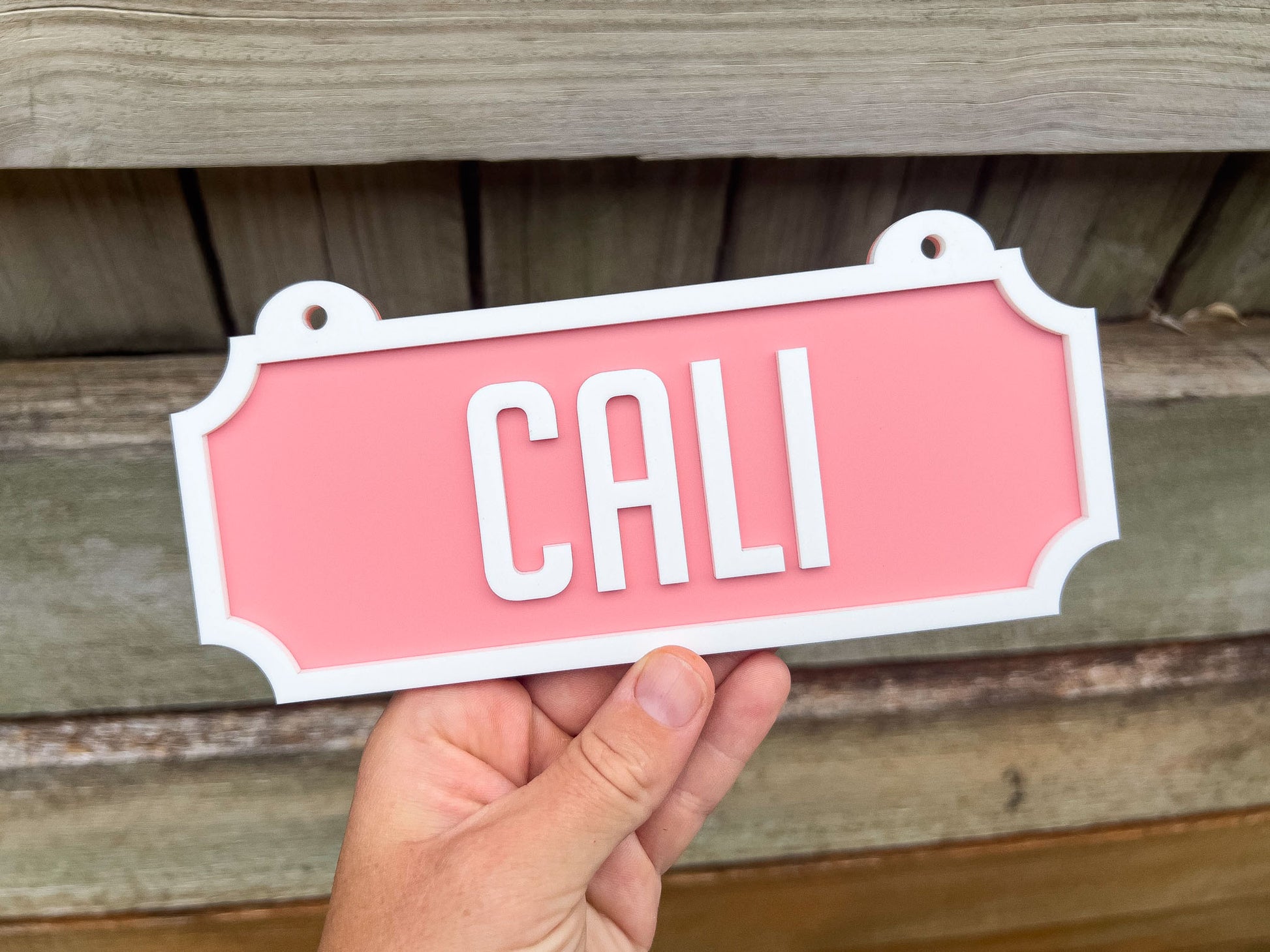 Hand holding a pink sign with 'CALI' on a wooden background | Coji Designs