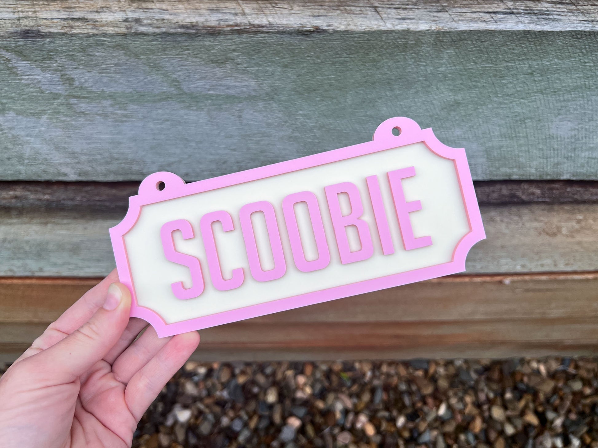 Hand holding a 'SCOOBIE' sign with pink border against a wooden background  | Coji Designs