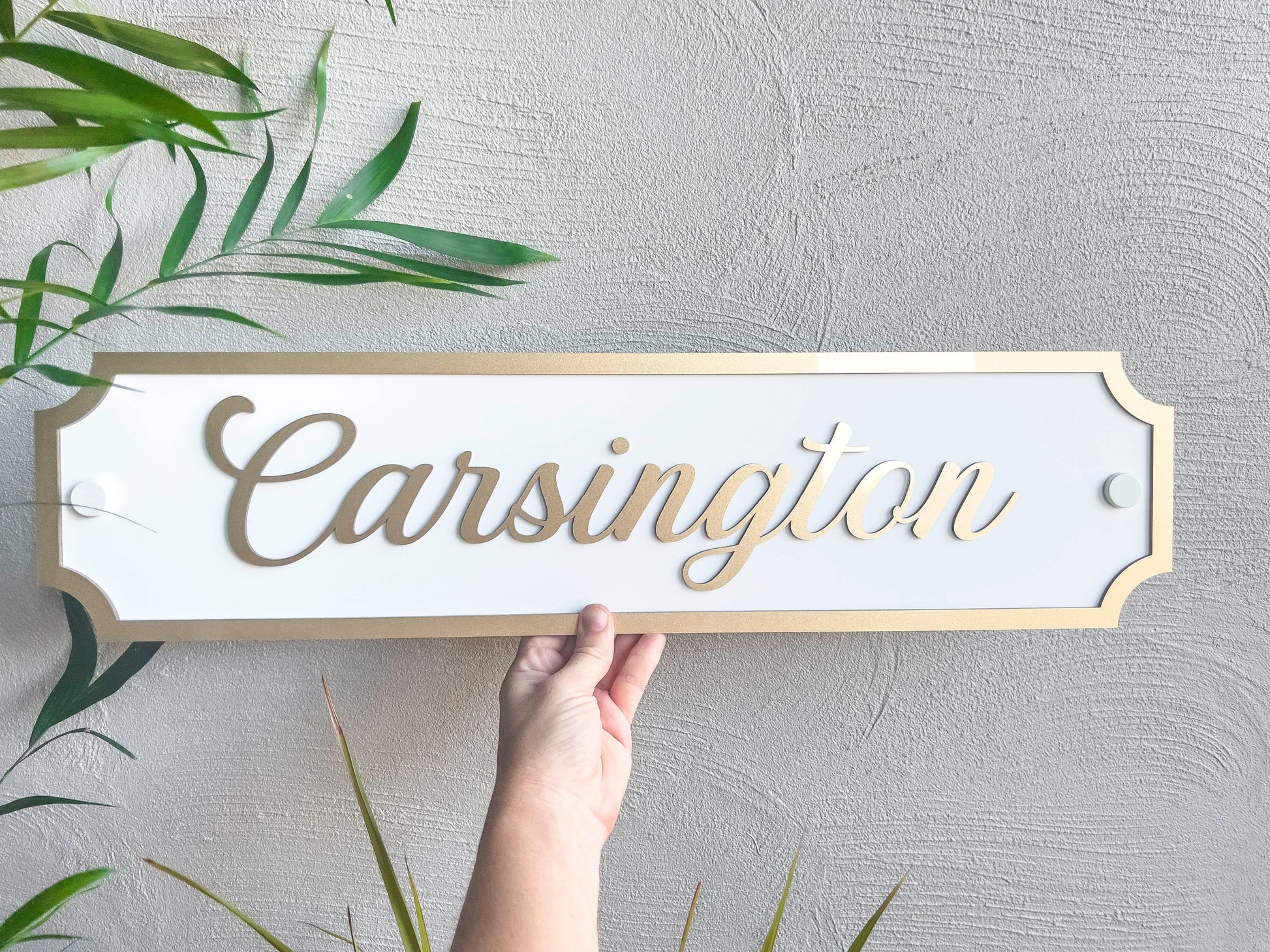 A stunning metallic gold and white tropical 3D acrylic panel that reads 'Carsington' in metallic gold and white held next to a palm tree.