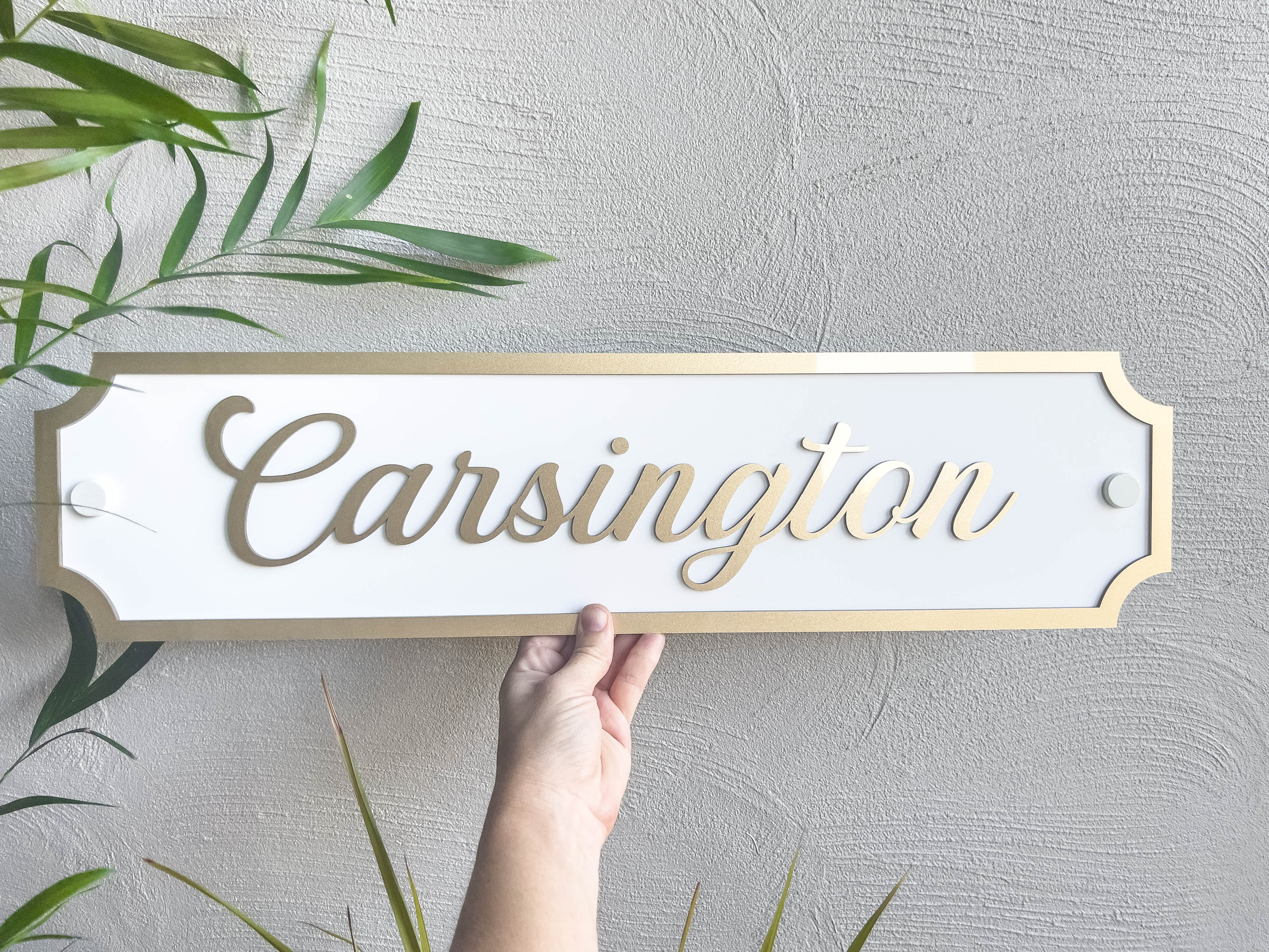 A stunning metallic gold and white tropical 3D acrylic panel that reads 'Carsington' in metallic gold and white held next to a palm tree.