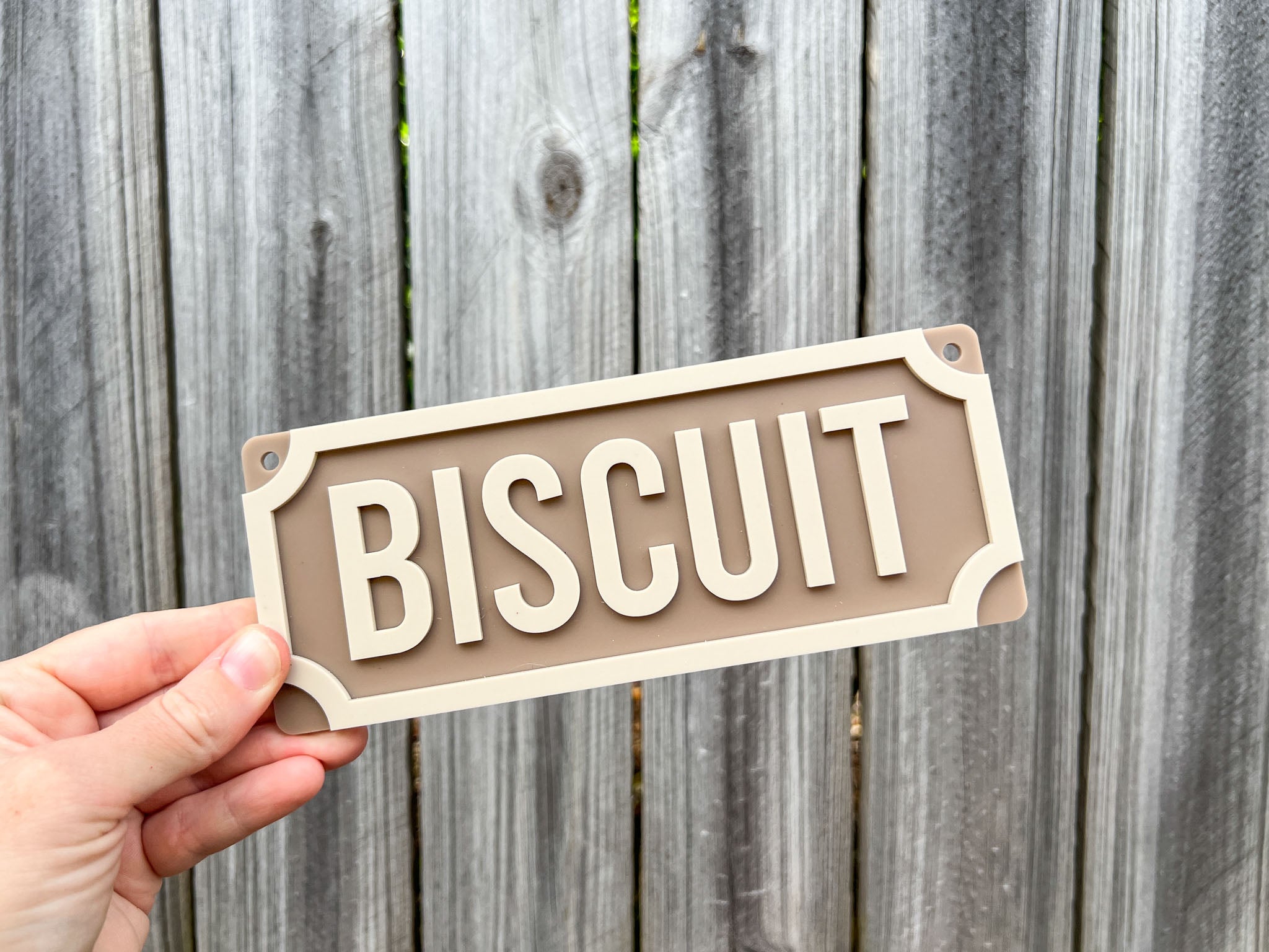Hand holding a rectangular sign with 'Biscuit' text against a wooden fence background