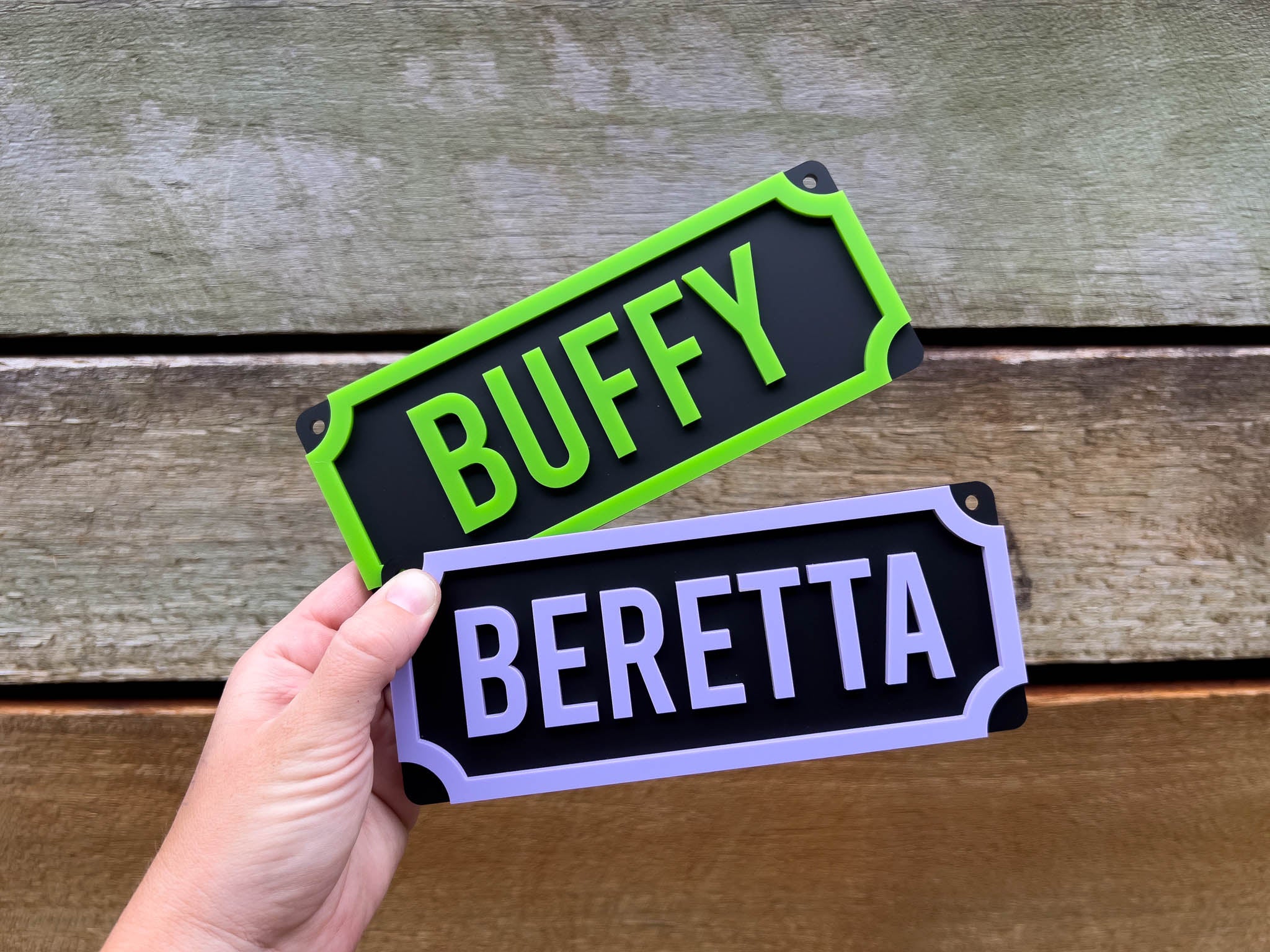Two nameplates with 'BUFFY' and 'BERETTA' text held against a wooden background.